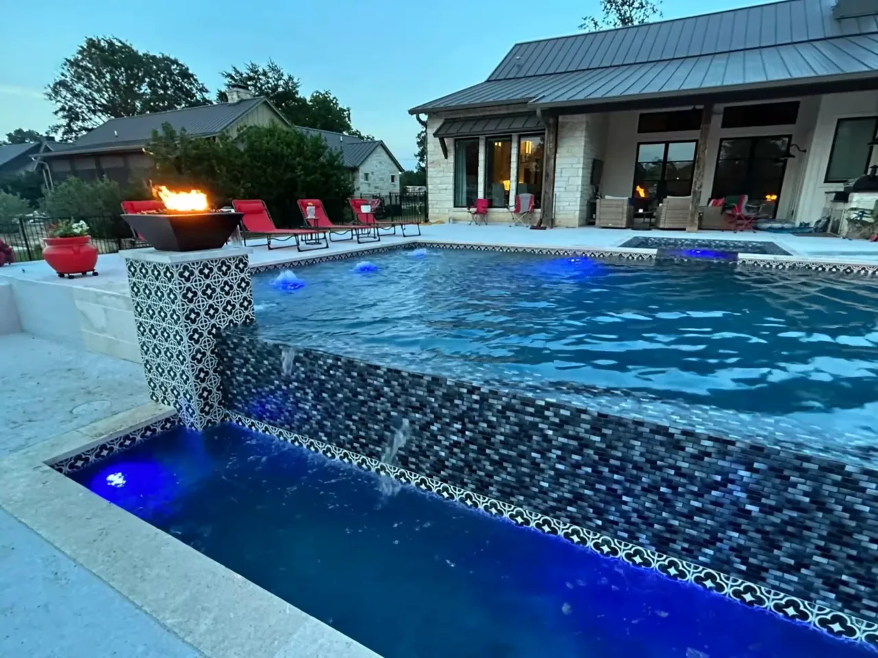 The Rivers Edge Pools hero loop, held still &mdash; a fire bowl and sheer-descent spillway on a custom gunite pool at dusk.