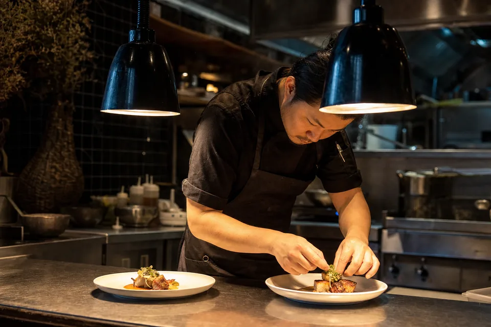 A chef plating two dishes under the pass lights on the kitchen line